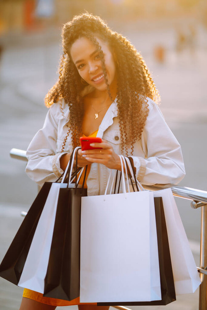 Giovane donna con borse della spesa utilizzando il telefono cellulare che cammina per strada. Stile Primavera. Consumismo, vendita, acquisti, shopping, lifestyle concept. - Foto, immagini