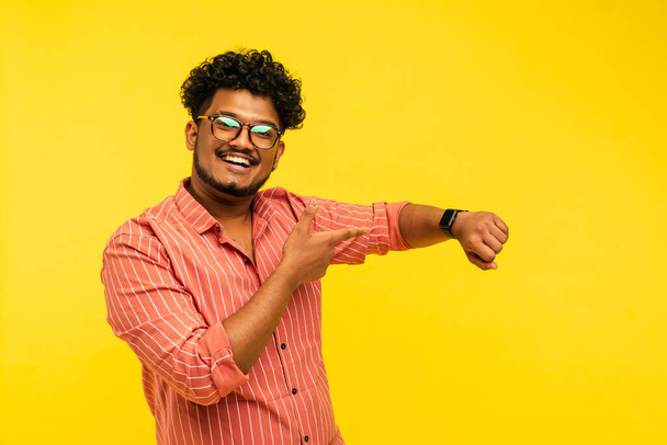 Hombre indio sonriente mostrando reloj en su mano, fondo amarillo con lugar para el texto. - Foto, imagen