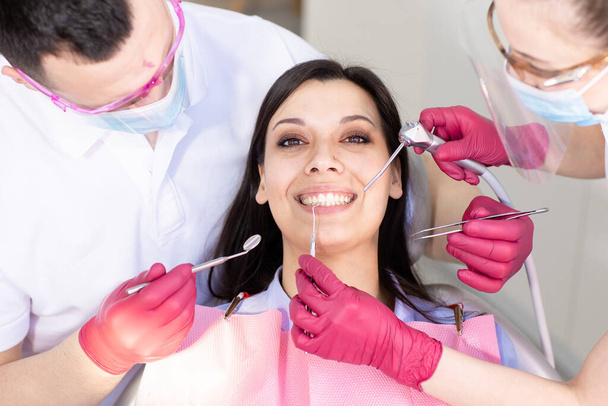 Portrait en gros plan d'une jeune femme souriante dans une chaise dentaire. Deux dentistes vérifient les dents et font leur hygiène. Soins de santé, médecine - Photo, image
