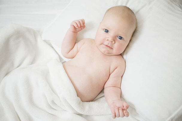 Naked newborn baby with a positive funny expression. Lies in bed on a pillow, covered with a white blanket. Caucasian, plump, cute baby. Eye contact. The concept of joy to have child, healthy toddler, good morning. - Foto, Imagen