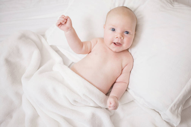 Naked newborn baby with a positive funny expression. Lies in bed on a pillow, covered with a white blanket. Caucasian, plump, cute baby. Eye contact. The concept of joy to have child, healthy toddler, good morning. - Fotografie, Obrázek