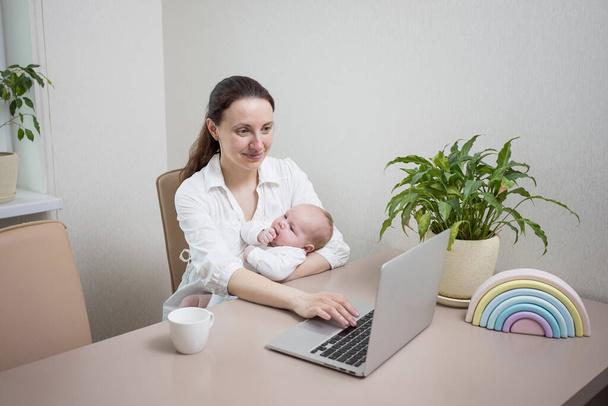 Mom with a newborn baby is working from home, typing on a laptop keyboard in the kitchen. Concept for online work, online shopping, busy mom, online consultation, new life, mother on maternity leave. - Photo, Image