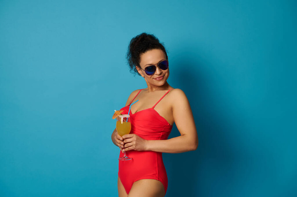 Приваблива жінка в червоному купальнику і сонцезахисних окулярах, дивлячись вниз з коктейлем в руках, стоїть ізольовано на синьому фоні з місцем для тексту
 - Фото, зображення