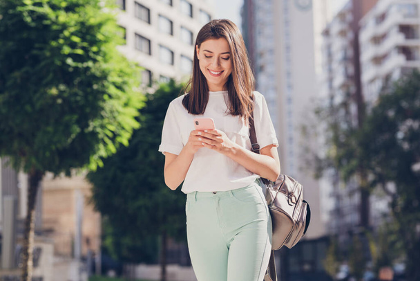 Photo portrait of female student with backpack typing sms using mobile phone walking along city street to college - Φωτογραφία, εικόνα