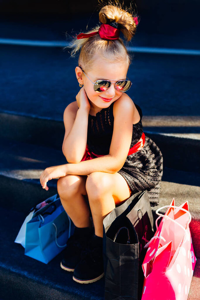 Bevásárlónap boldog gyerekkel. mosolygó kislány napszemüvegben ül a lépcsőn a csomagok mellett.. - Fotó, kép