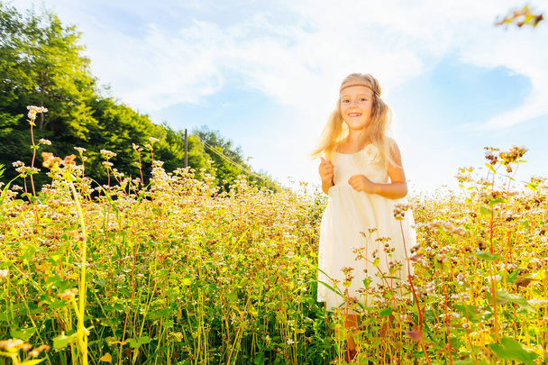 Felice bambina carina in un vestito bianco in un campo con fiori al tramonto estivo. Il concetto di gioia di vita, felicità, successo - Foto, immagini