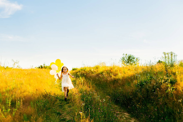 Beyaz elbiseli güzel bir kız yeşil çimlerde koşuyor ve elinde balonlar tutuyor.. - Fotoğraf, Görsel