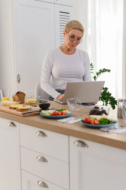 Idősebb nő a laptopnál dolgozik a konyhában. Úgy néz ki, főzés receptek online. - Fotó, kép