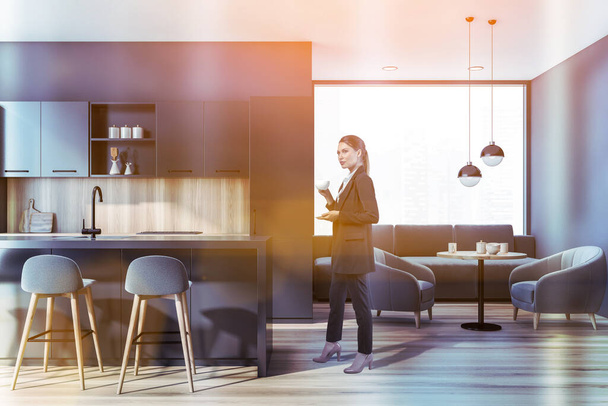 Side view woman at kitchen room with furniture and panoramic window, lens flare. Businesswoman drinking coffee in luxury dining room, wooden floor, toned image - 写真・画像