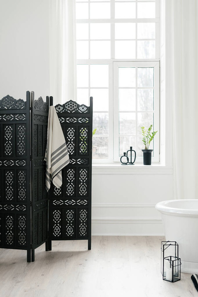 Κάθετη λήψη από μαύρη πτυσσόμενη οθόνη με κρεμάμενη πετσέτα κοντά σε μεγάλο παράθυρο στο μοντέρνο και ελαφρύ μπάνιο. Έννοια της επίπλωσης και διακόσμησης του σπιτιού στη σύγχρονη διακόσμηση εσωτερικών χώρων - Φωτογραφία, εικόνα