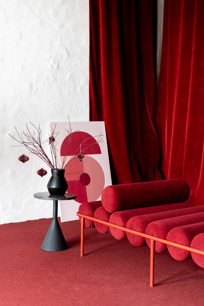 Japanisches Interieur für den Ruhebereich. Rotes, einzigartiges Sofa mit zylindrischen Kissen und Velourvorhängen, Äste in der Vase auf minimalistischem Teetisch und abstrakte Malerei auf dem Boden mit weichem Teppich - Foto, Bild