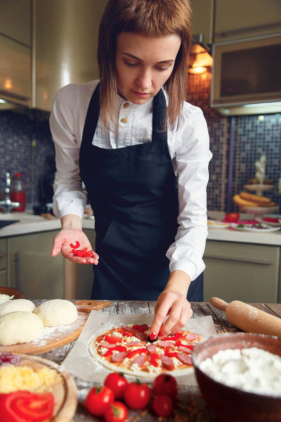 Woman in white shirt and apron putting the ingredients on the pizza. Pizza concept. Production and delivery of food - Zdjęcie, obraz