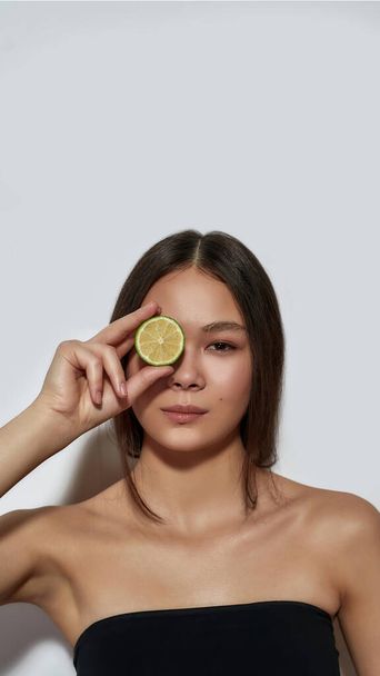 Cute young asian girl holding lime slice near eye - Foto, afbeelding