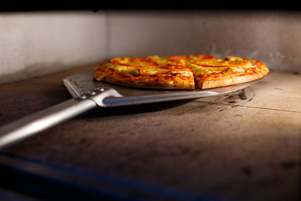 Heiße leckere amerikanische Pizza mit Tomatenkäse und Fleisch mit dicker Kruste auf einer Metallschaufel, der Bäcker holt die Pizza aus dem Ofen - Foto, Bild