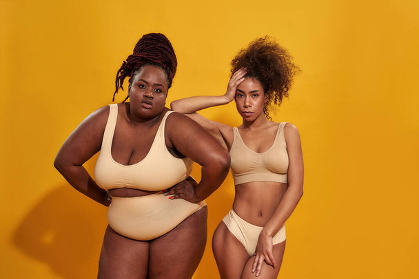 Studio shot of two confident plus size and slim african american female models in beige underwear looking at camera while posing together isolated over yellow background - Foto, immagini