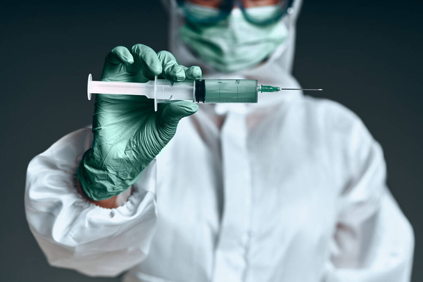 Protection against infection. The doctor is dressed in a protective suit, protective glasses and a protective mask holds a syringe with a vaccine in his hands. Isolated on a dark background. Selective focus - Foto, imagen