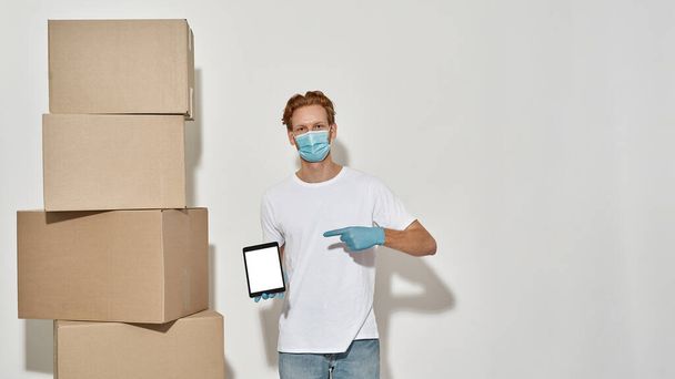 Young deliveryman stands near cardboard boxes pointing at tablet - Foto, Bild