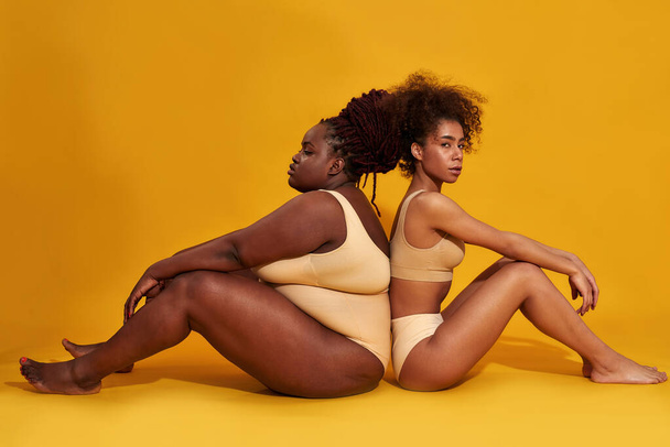 Full length shot of two diverse african american female models in underwear sitting together isolated over yellow background - Photo, Image