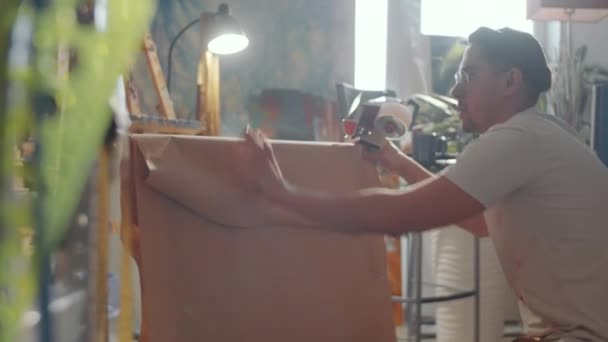 Професійний художник-чоловік використовує дозатор клейкої стрічки під час упаковки картини в крафт-папері перед відправкою
 - Кадри, відео