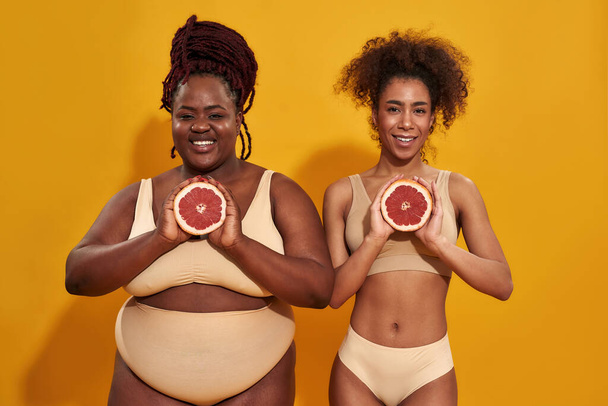 Studio shot of two joyful african american women in underwear smiling at camera, holding halfs of juicy grapefruit in front of them, posing isolated over orange background - Photo, image