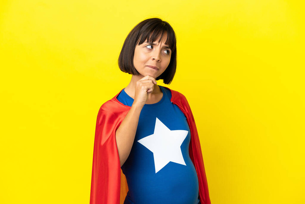 Superheldin schwangere Frau isoliert auf gelbem Hintergrund mit Zweifeln und Denken - Foto, Bild