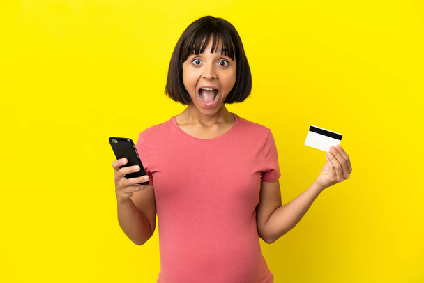 Nuori sekarotuinen raskaana oleva nainen, joka on eristetty keltaisella taustalla ostamalla matkapuhelimella ja pitämällä luottokorttia, jolla on yllättynyt ilme - Valokuva, kuva