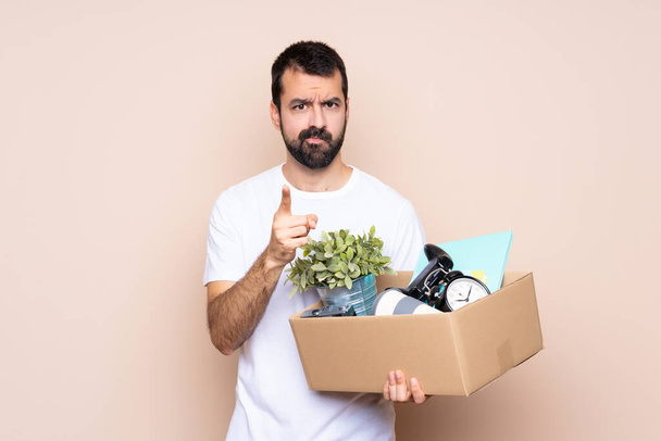 Man holding a box and moving in new home over isolated background frustrated and pointing to the front - Photo, Image