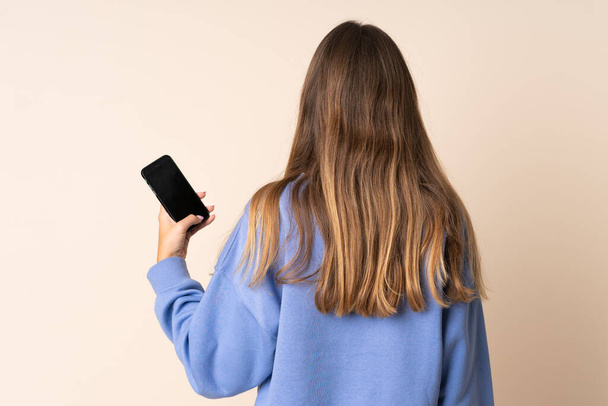 Young Lithuanian woman using mobile phone isolated on beige background in back position - 写真・画像