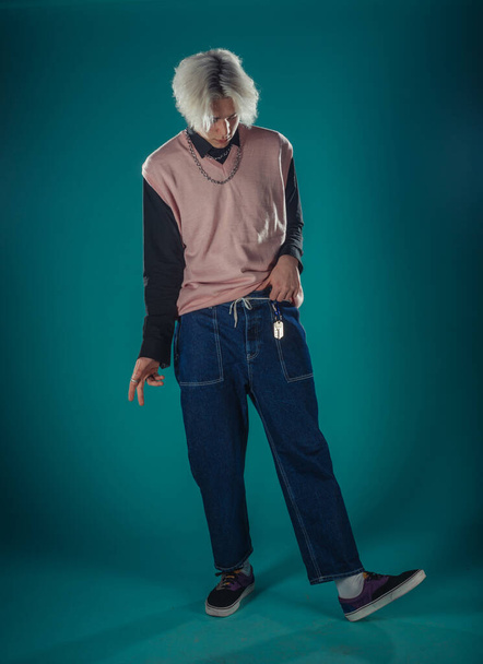 Young adult man in casual outfit wear looking at camera, holding skateboard near face and standing at studio on blue background - 写真・画像