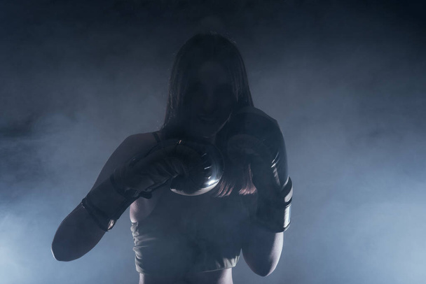 Dark contrast photo with smoke in the background of a focused strong woman with boxing gloves practicing punches - Foto, afbeelding