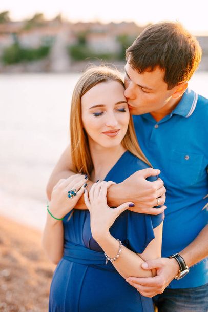 Man gently kisses smiling pregnant woman in a blue dress hugging her shoulders - Photo, Image