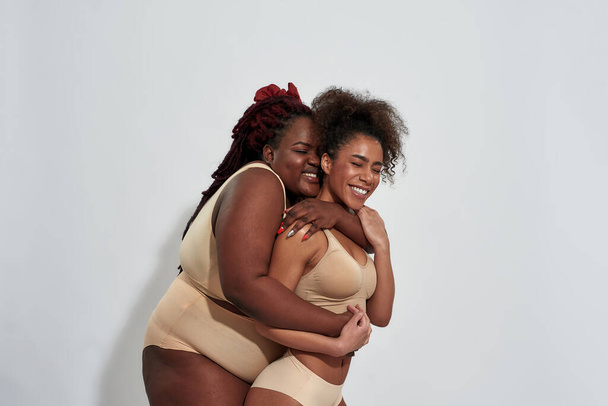 Two friends, african american women in underwear with different body shape embracing and smiling with eyes closed, posing isolated over gray background - Fotografie, Obrázek