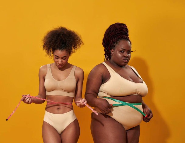Two african american women with different body shape measuring their waist using metric tape measure, standing together isolated over orange background - Foto, Imagen