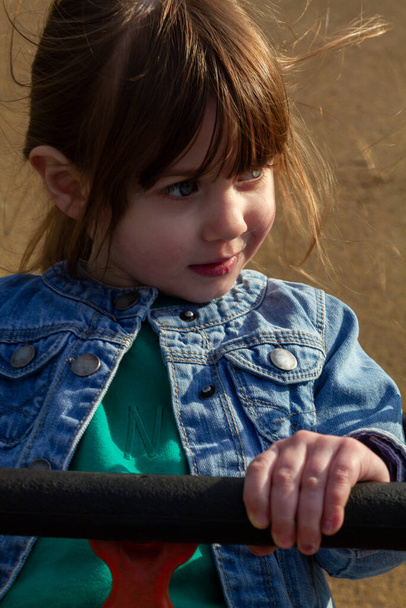 A cute, brown-haired, blue-eyed girl wearing a blue jacket and green jumper sitting in a swing on a sunny spring day - Photo, Image