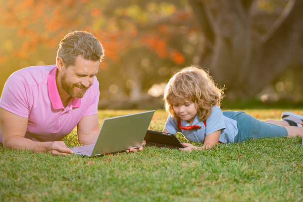 Portrait of man with son using digital tablet outside. New normal back to school. Elementary scholar pupil sitting with teacher on grass. - Fotoğraf, Görsel