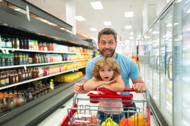 Happy smiling father and child son with shopping cart buying food at grocery store or supermarket. - Foto, Imagem