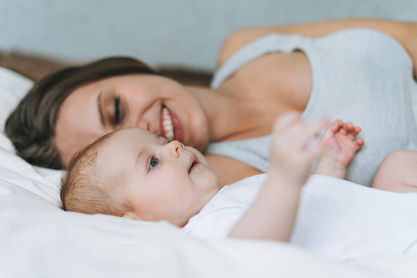 Jonge moeder hebben plezier met schattige baby meisje op het bed met wit linnen, natuurlijke tinten, liefde emotie - Foto, afbeelding