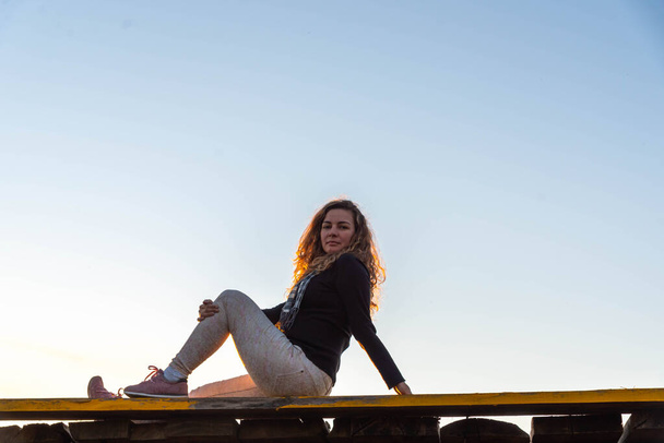 Dospělá žena sedí na dřevěném mostě na modrém přírodním pozadí. Žena pózující na focení za soumraku. Dramatické a barevné nebe. Venkovská krajina s lidmi na scéně. Přirozený životní styl. - Fotografie, Obrázek