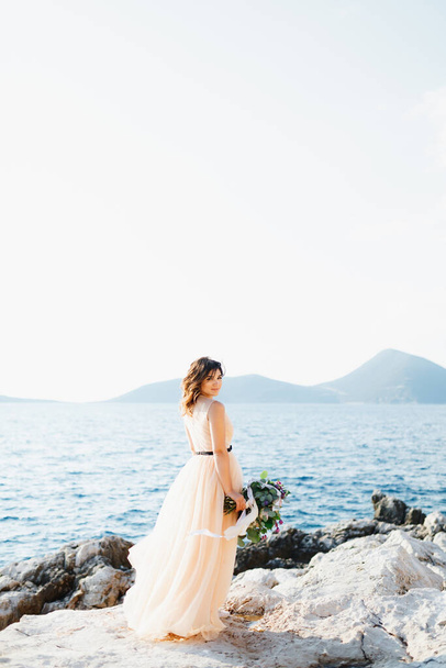 Lächelnde Braut im pastellfarbenen Brautkleid steht mit einem Blumenstrauß in der Hand auf einem Felsen über dem Meer - Foto, Bild