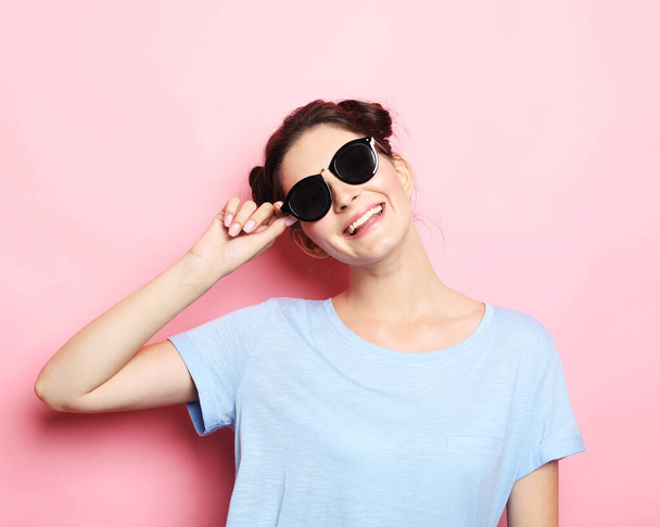 Happy time. Young cute smiling brunette woman wearing casual blue shirt and sunglasses posing over pink background. - Zdjęcie, obraz