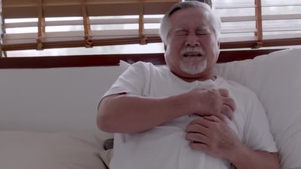 Ασιάτης ηλικιωμένος άνδρας με πονοκέφαλο κάθεται σε ένα κρεβάτι το πρωί - Πλάνα, βίντεο