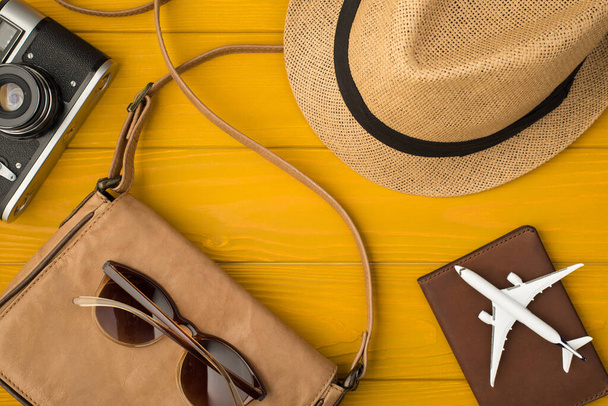 Güneş şapkası çantasının üstünde güneş gözlüğü pasaportu ve sarı ahşap arka planda izole edilmiş uçak resmi var. - Fotoğraf, Görsel