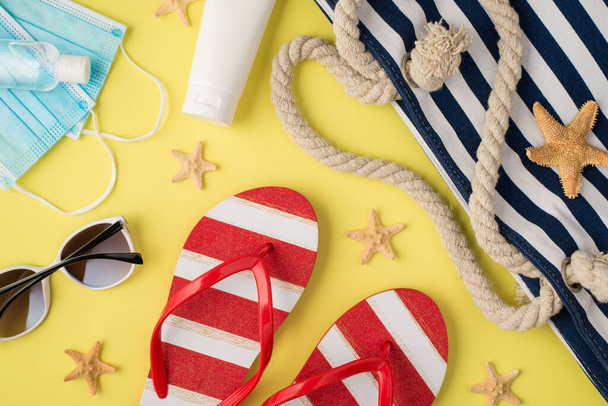 Top view photo of sanitizer medical masks cream bottle beach bag striped flip-flops sunglasses and starfishes on isolated pastel yellow background - Fotó, kép