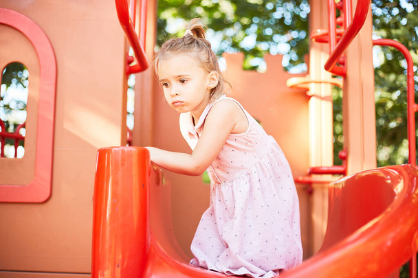 Cute caucasian girl boring play alone in play outdoor park in summer. Emotional funny girl playing at the playground.  - Photo, image