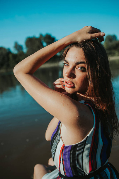 A portrait of a confident brunette woman wearing a striped dress and posing near a lake in a park - Fotografie, Obrázek