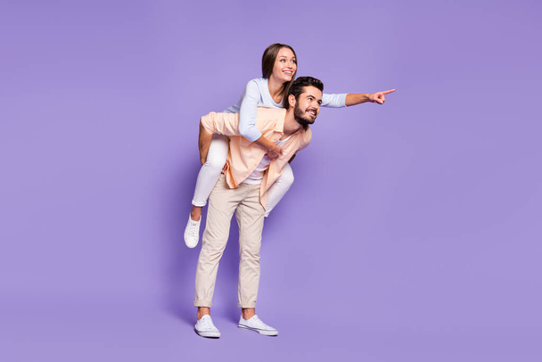 Full size photo of happy young couple man piggyback woman point finger empty space isolated on purple color background - Foto, Bild