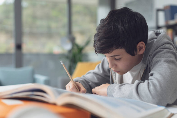 Lindo chico sentado en el escritorio y estudiando, está leyendo y escribiendo notas - Foto, Imagen