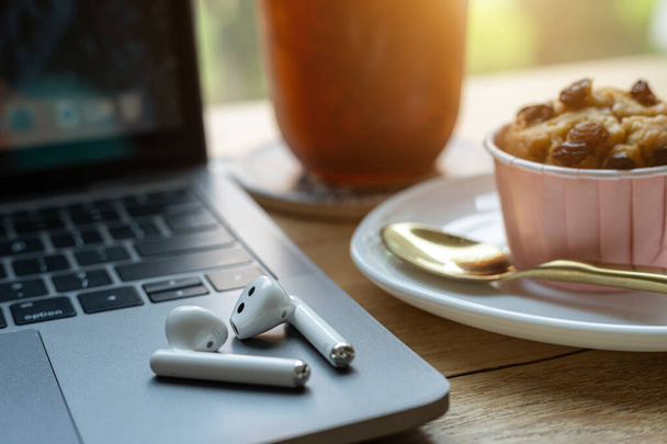 Close-up of Homemade Banana cup cake and ice coffee in cup mug with wireless earphones on laptop computer on wood desk office desk in coffee shop at the cafe,during business work concept - Foto, Imagem