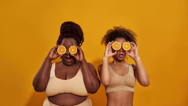 Dwie afrykańskie kobiety w bieliźnie zasłaniające oczy połówkami dojrzałej soczystej pomarańczy, pozujące razem na pomarańczowym tle - Zdjęcie, obraz