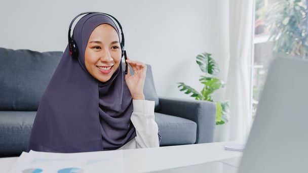 Asia muslim lady wear headphone using laptop talk to colleagues about plan in conference video call while working from home at living room. Social distancing, quarantine for corona virus prevention. - Photo, image